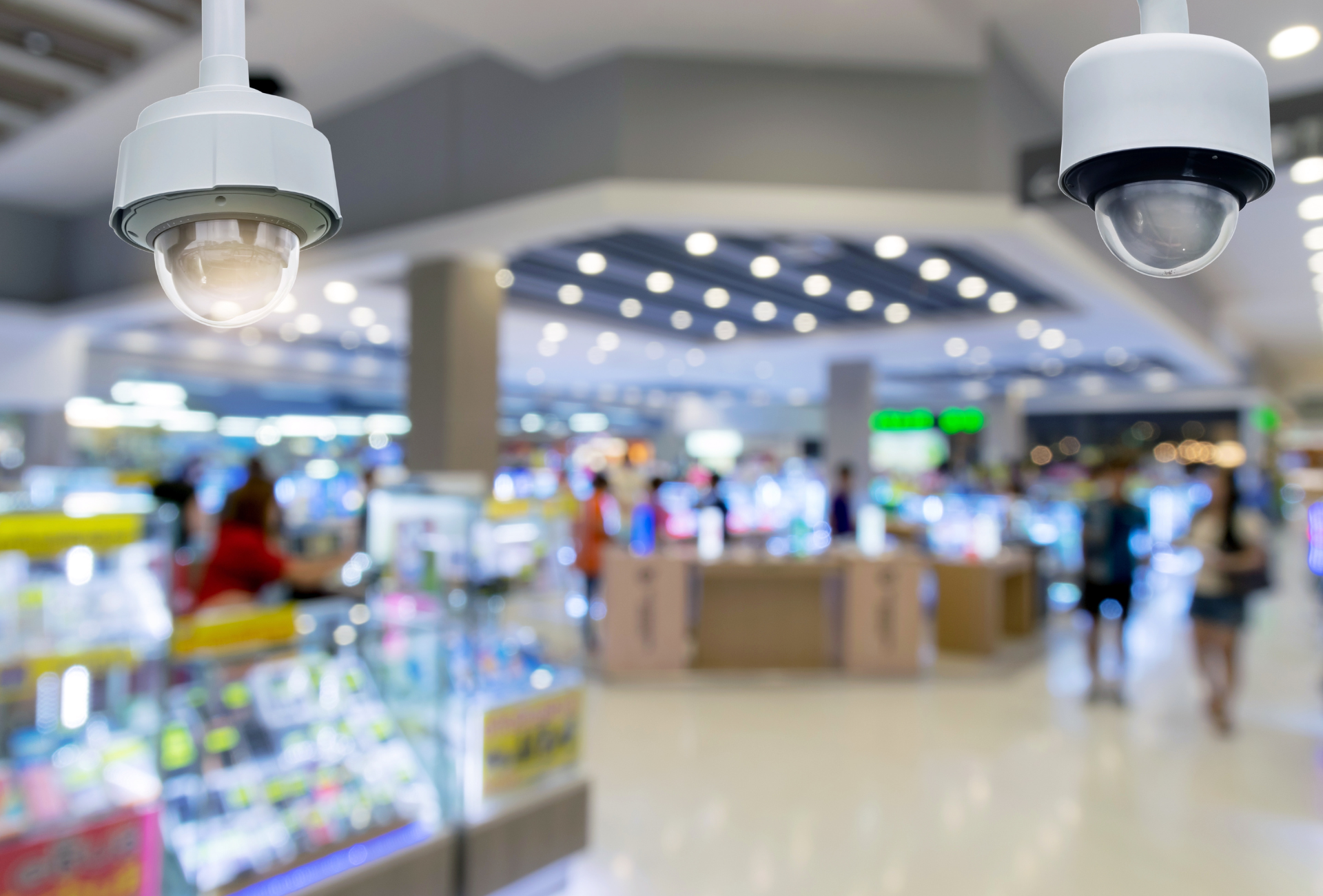 Two security cameras are hanging from the ceiling of a shopping mall.